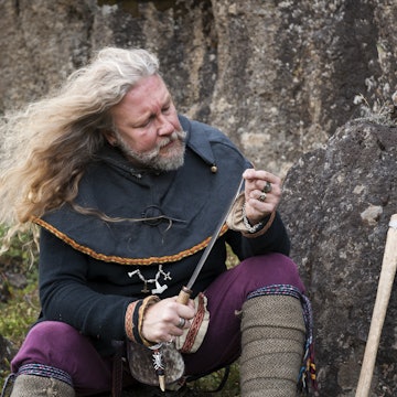 Blond Viking male reenactor in full warrior armour and battle gear is sitting on a rock cleaning his knife in the historic location where Vikings assembled annually to recite and discuss laws, Thingvellir National Park, Pingvellir, Iceland