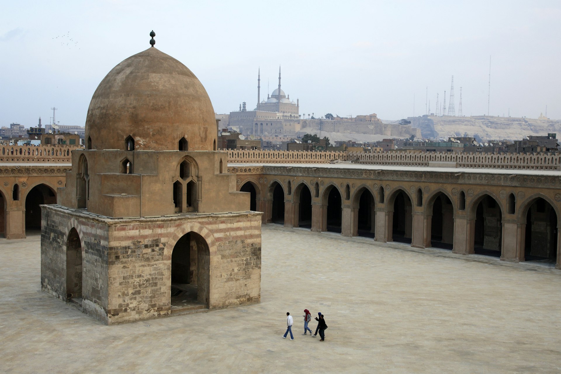 Four people walk through a courtyard with a large domed structure and arched cloisters