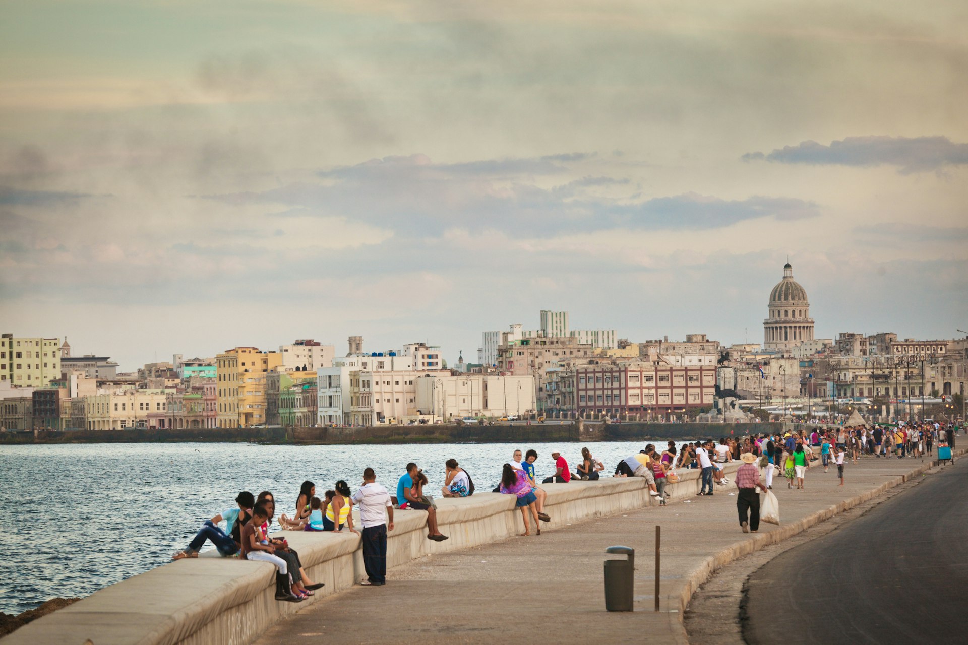 People sit along a sea wall in the evening as the sun starts to set. The city scape, dominated by a large domed building, stretches out behind them