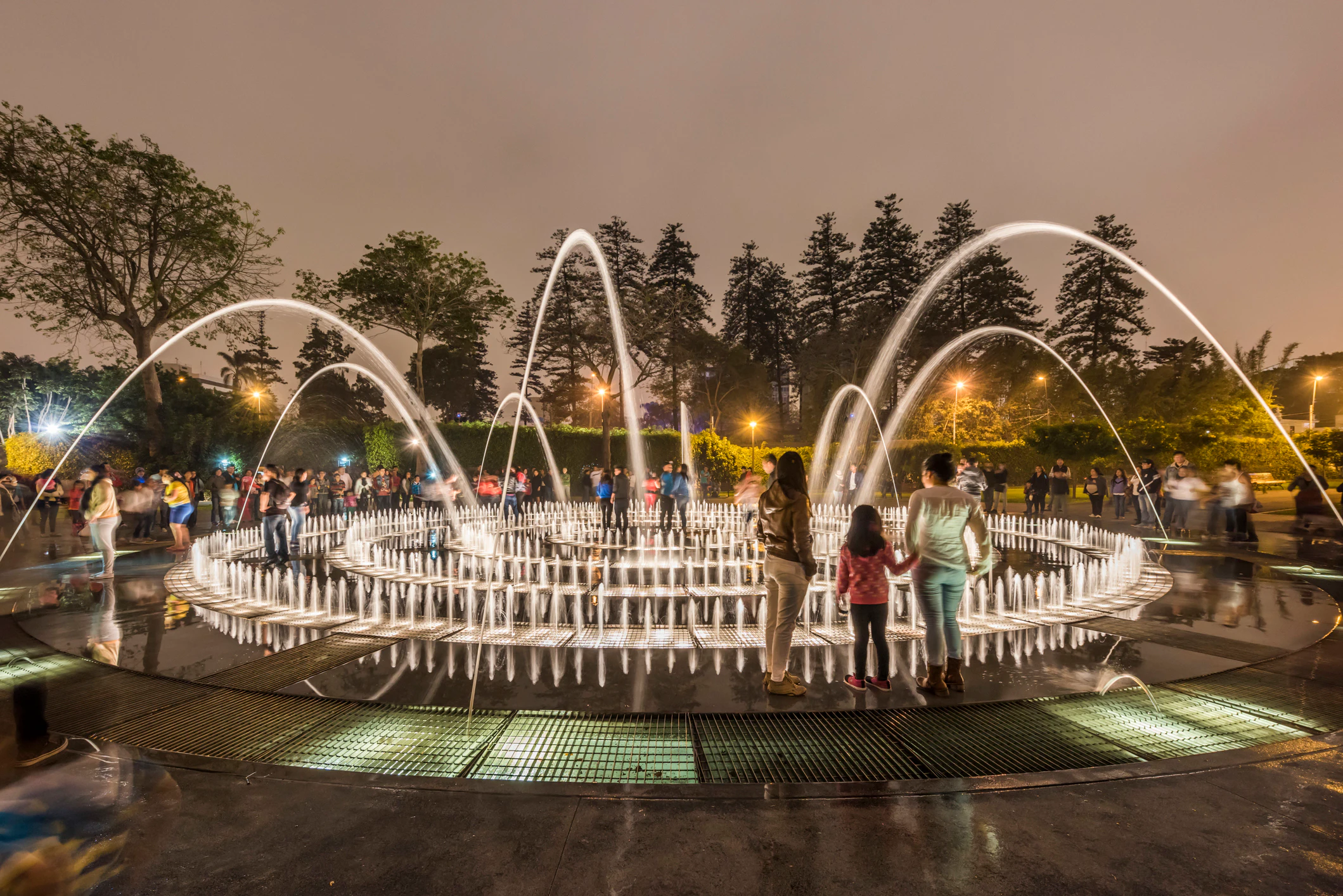 People stand in a park by an elaborate fountain with dozens of jets.