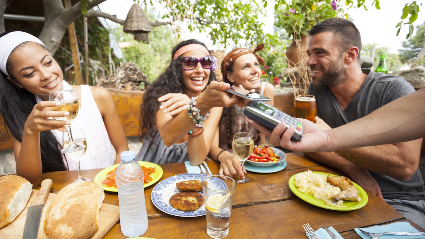 Young people using smart phone for contactless payment in restaurant - stock photo
Young people having fun on outdoor restaurant. Food and drinks on table. One woman with sunglasses using smart phone for contactless payment. On foreground male hand holding credit card reader © portishead1 / Getty