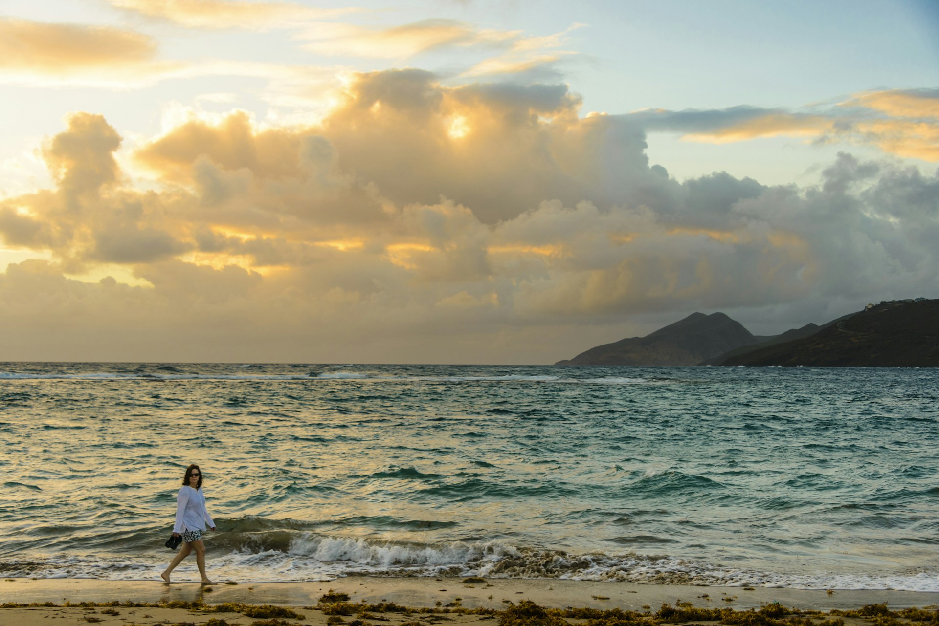 A barefoot woman walks along the shore of a sandy beach as the sun rises