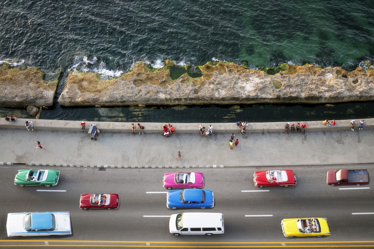 Large group of vintage American cars speeding along the Malecon in Havana, Cuba, motion blur, people sitting on sea wall, Caribbean Sea is visible in the background, elevated view.
993970062