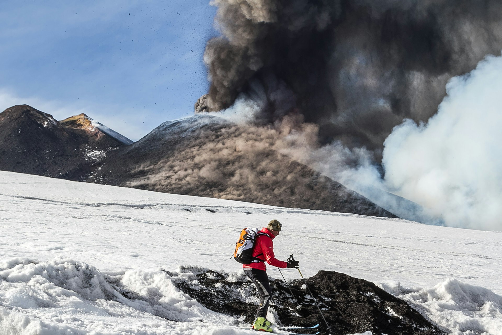 A skier heads down a mountain as a nearby volcano belches soot and black clouds into the sky 