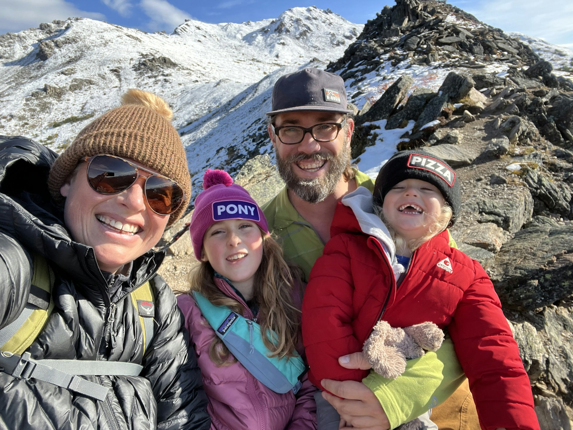 Uma família sorridente (dois adultos e duas crianças) no topo da Savage Alpine Trail em Denali com as montanhas cobertas de neve ao fundo