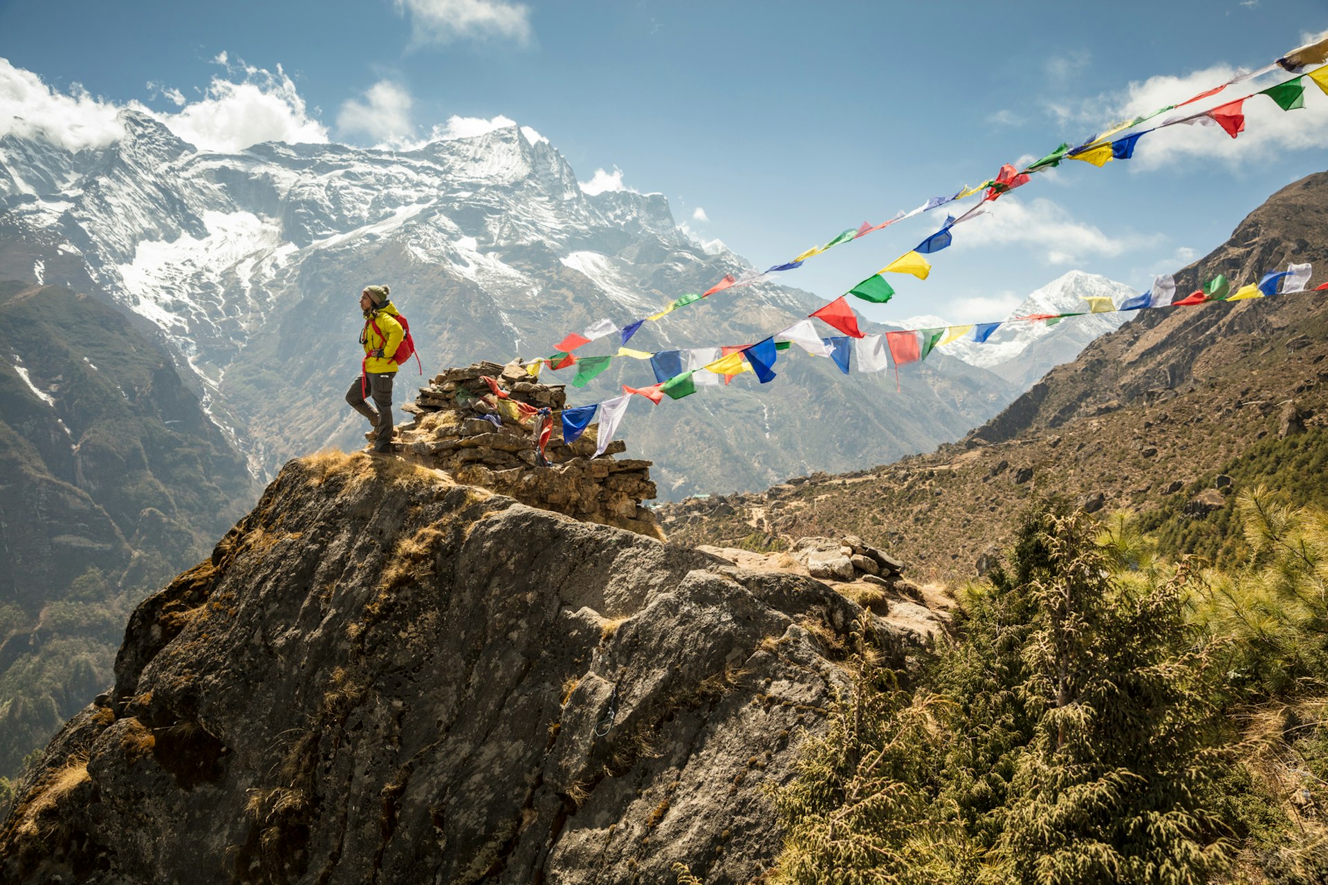 A hiker stands at the top of a tall mountain in Nepal with brightly-colored prayer flags flapping in the wind