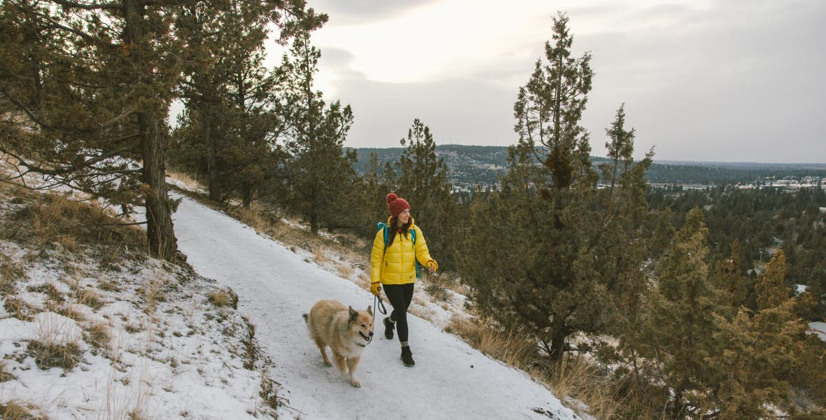 Woman in yellow coat walking with dog along snowy trail