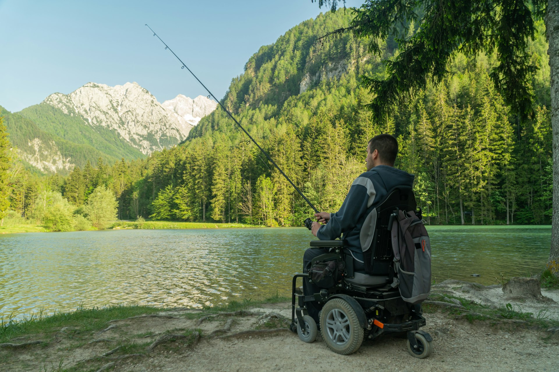 Young man in a wheelchair fishing at the beautiful lake on a sunny day, with mountains in the back 