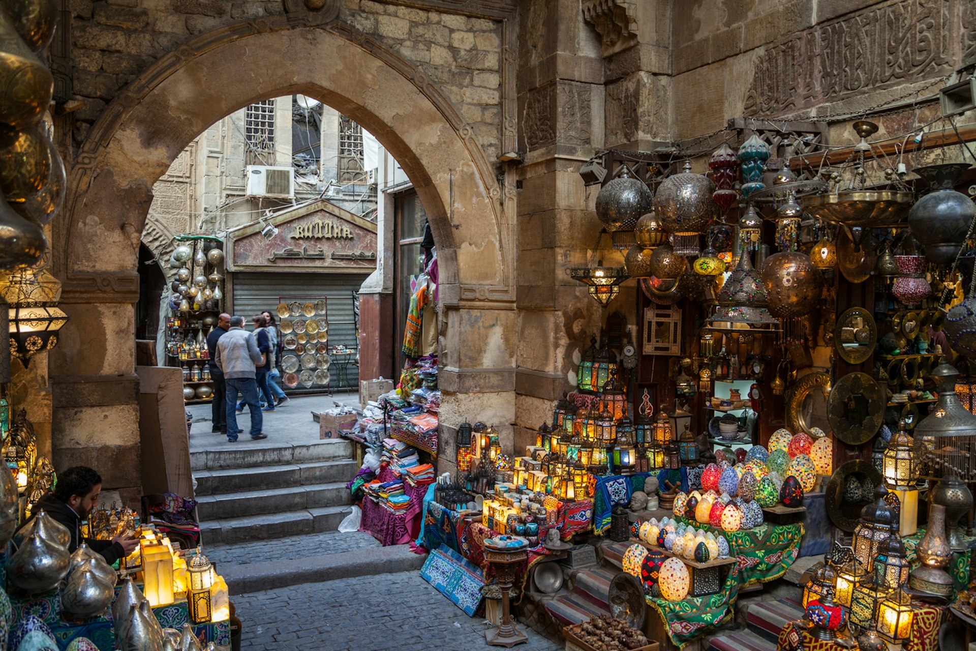 A stall packed with lamps and lanterns in a souk with another pathway leading to more stalls through an arched doorway