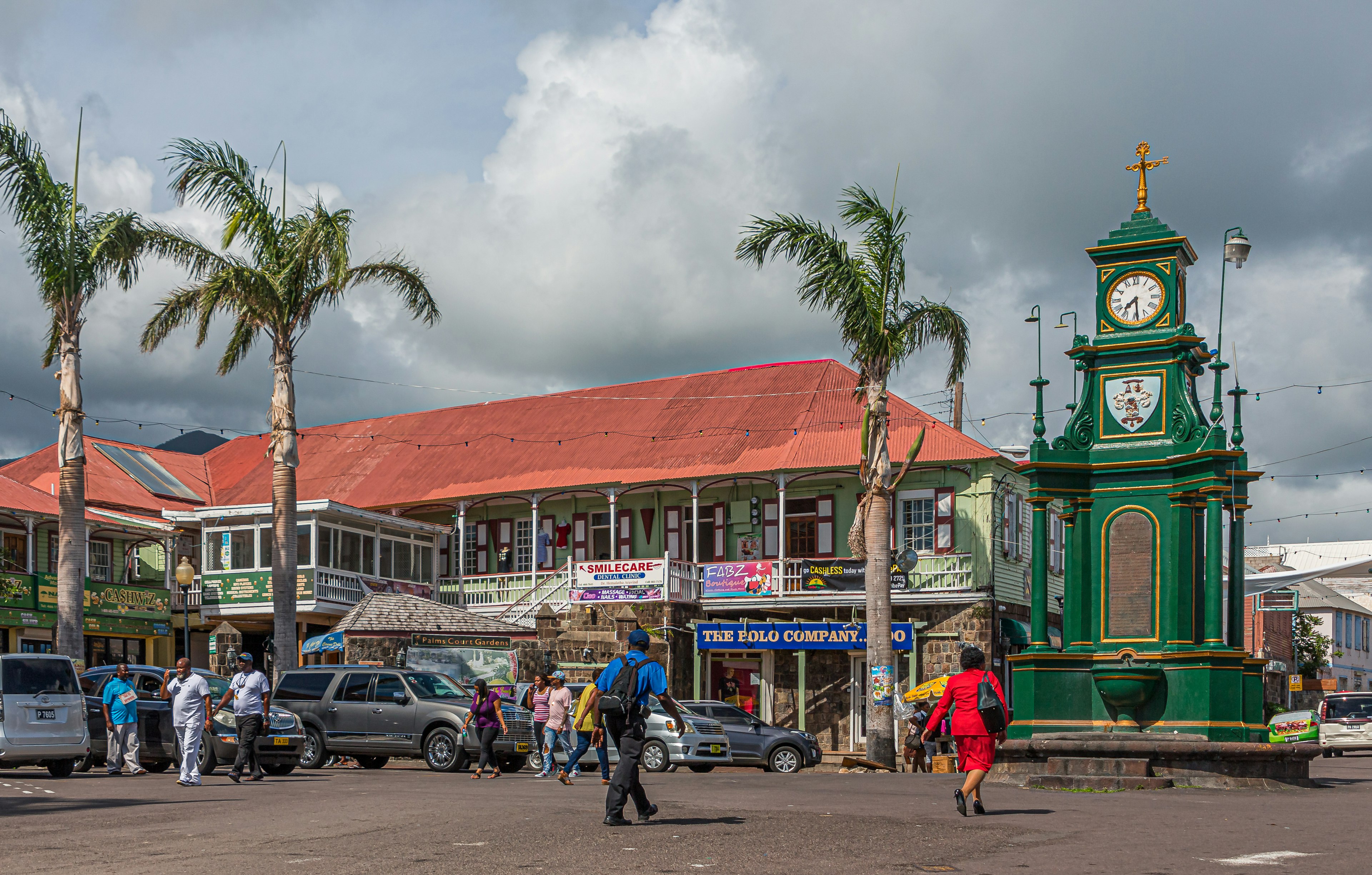 A small green clock tower stands in the center of a road junction in a town