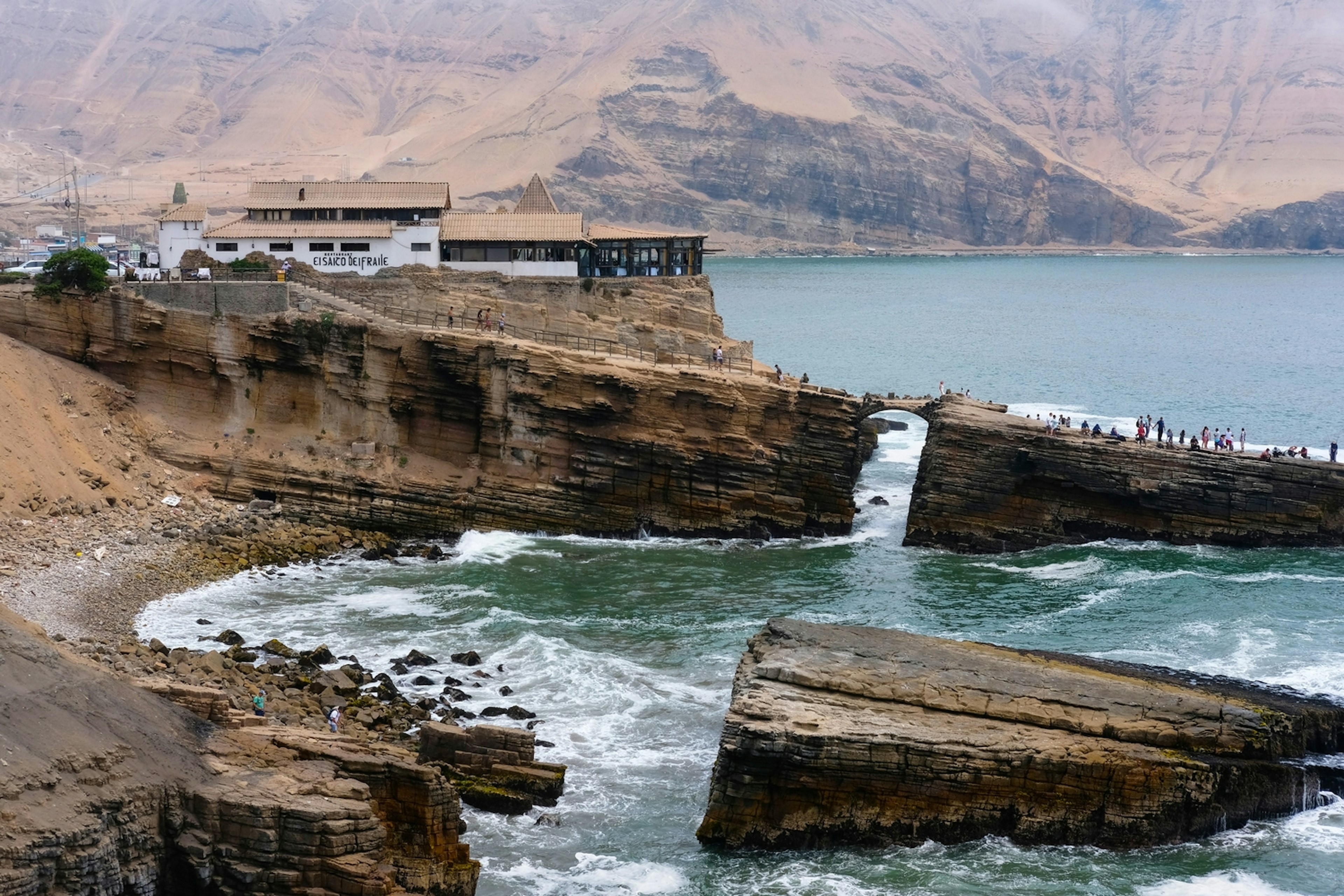 A rocky outcrop stretches out to sea. At the central point is a gap where the sea flows through and a narrow bridge above