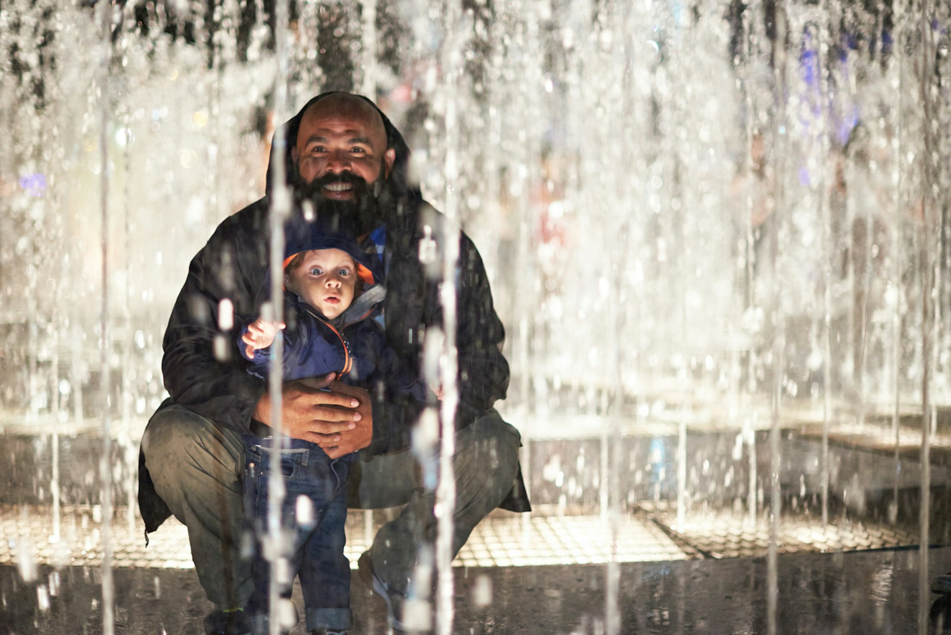 A father sits with a toddler in among the jets of a fountain. The toddler is reaching out at the water.