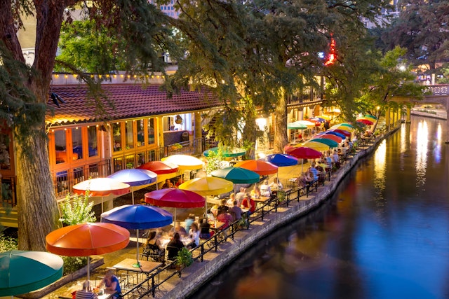 Colorful umbrellas line a waterway with people dining nearby