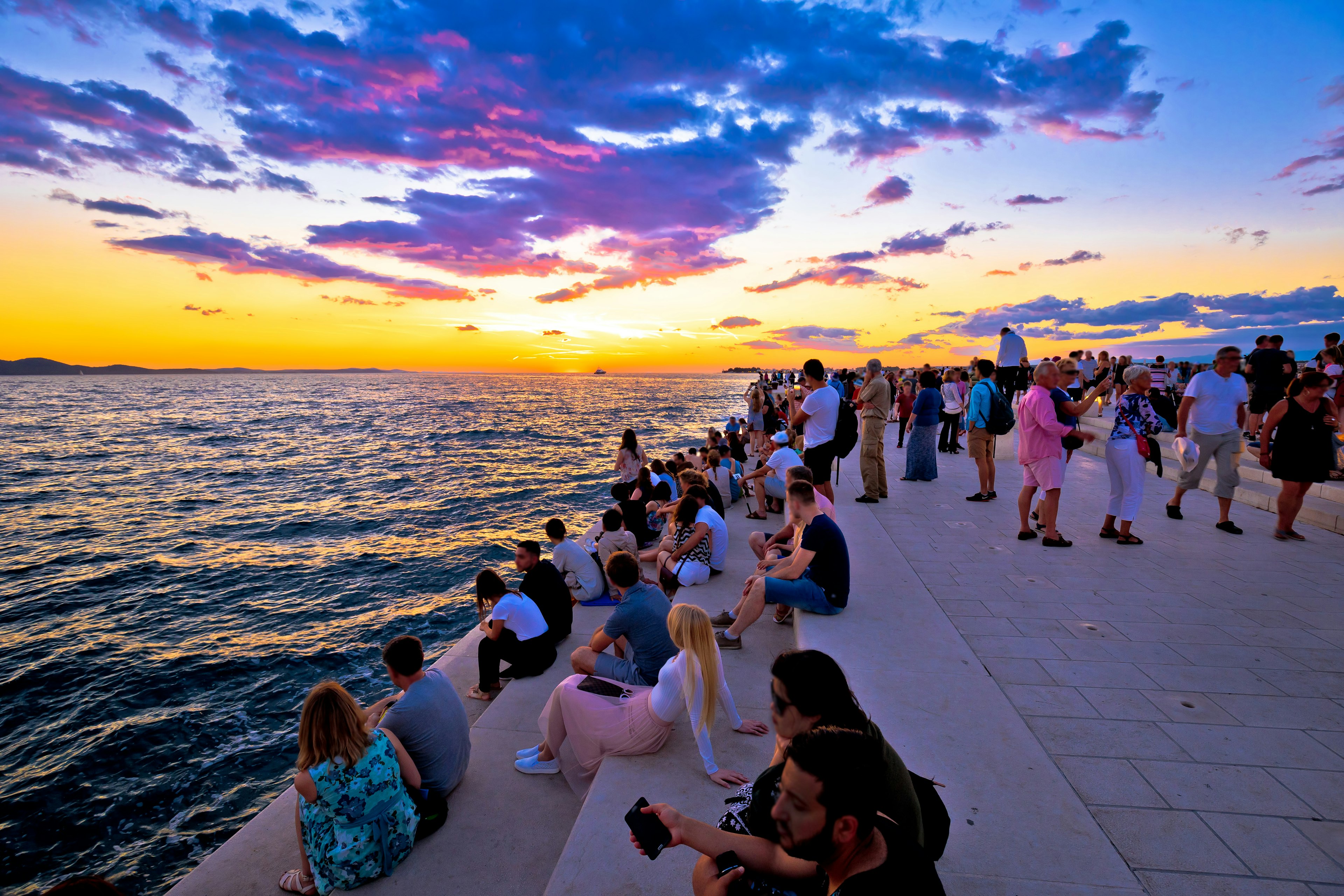 People sit on steps by the sea as the sun casts oranges, yellows, blues and purples across the sky at sunset