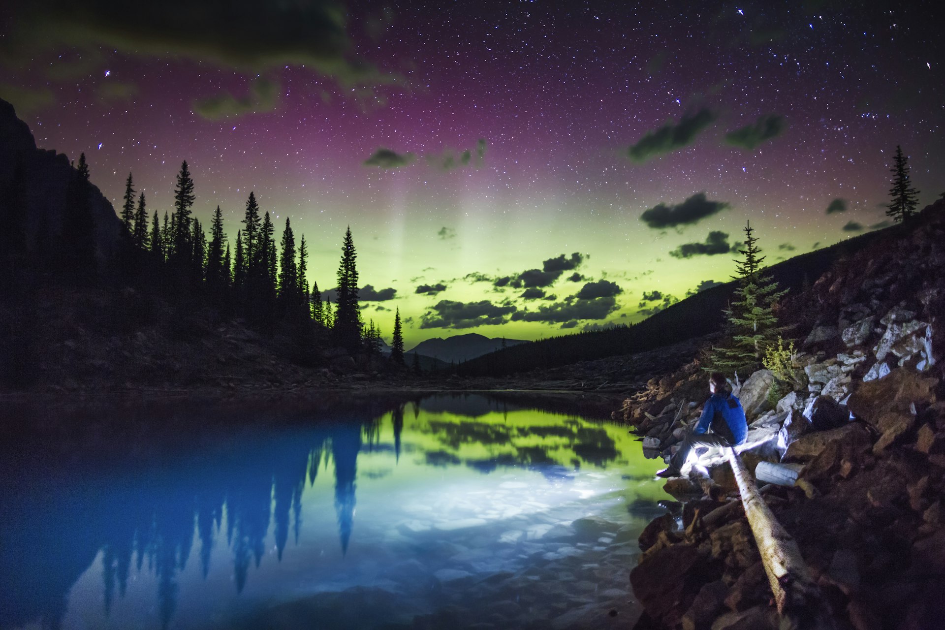 A person sits by a calm lake at night, with the Northern Lights (aurora borealis) shimmering in the sky above, reflecting in the water, surrounded by silhouetted pine trees.