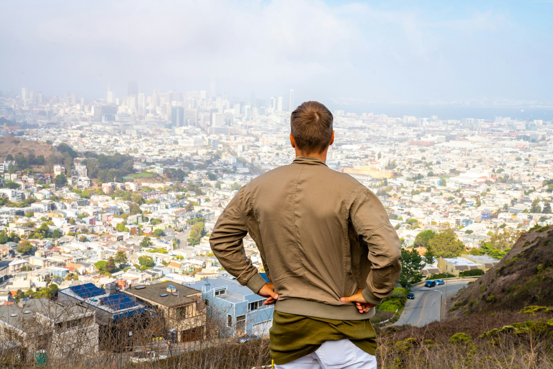 A man with his back to the camera stands at a high vantage point, hands on hips, overlooking a panoramic view of San Francisco)