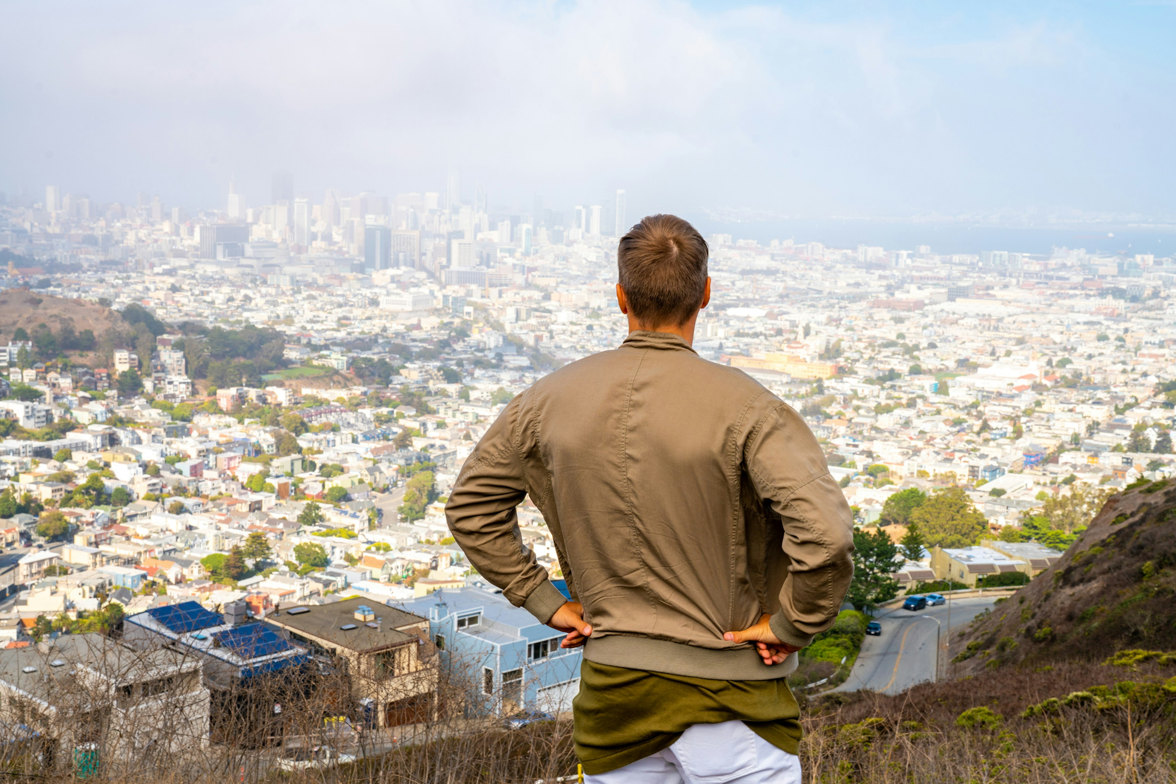 A man with his back to the camera stands at a high vantage point, hands on hips, overlooking a panoramic view of San Francisco)