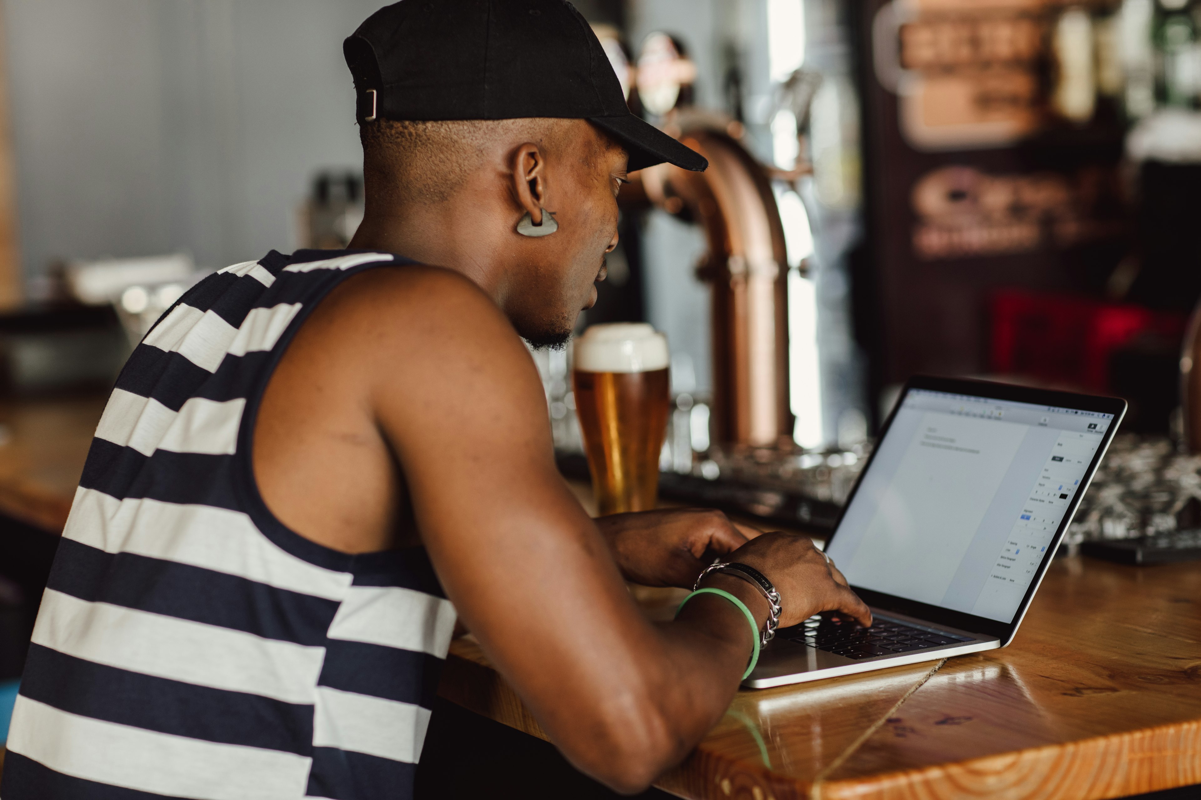 A man is working on a laptop at a bar counter, with a pint of beer next to him.