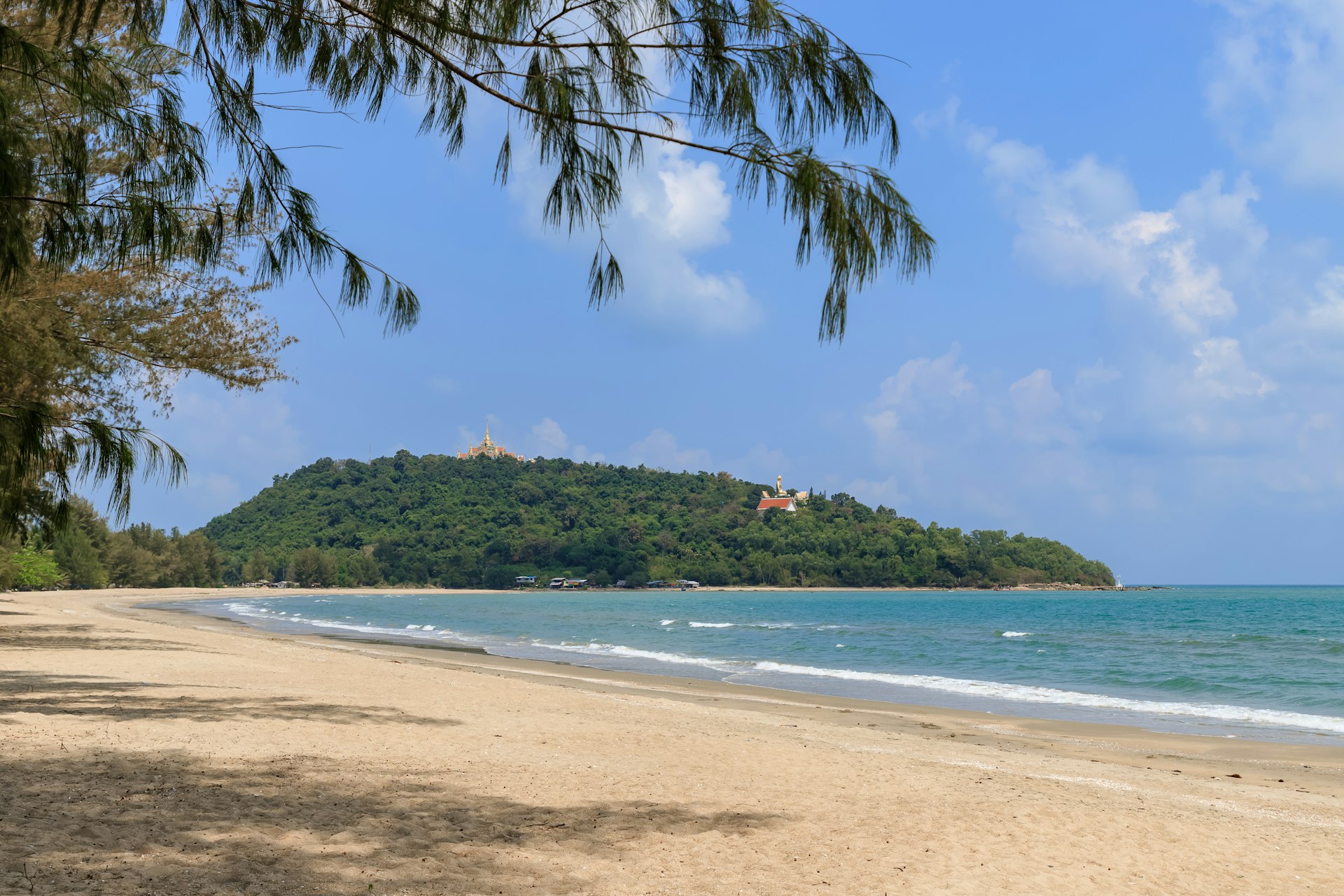 A beach landscape showing a forested island with a prominent building at the top of a hill, viewed from a sandy shore under the shade of an overhanging tree branch.