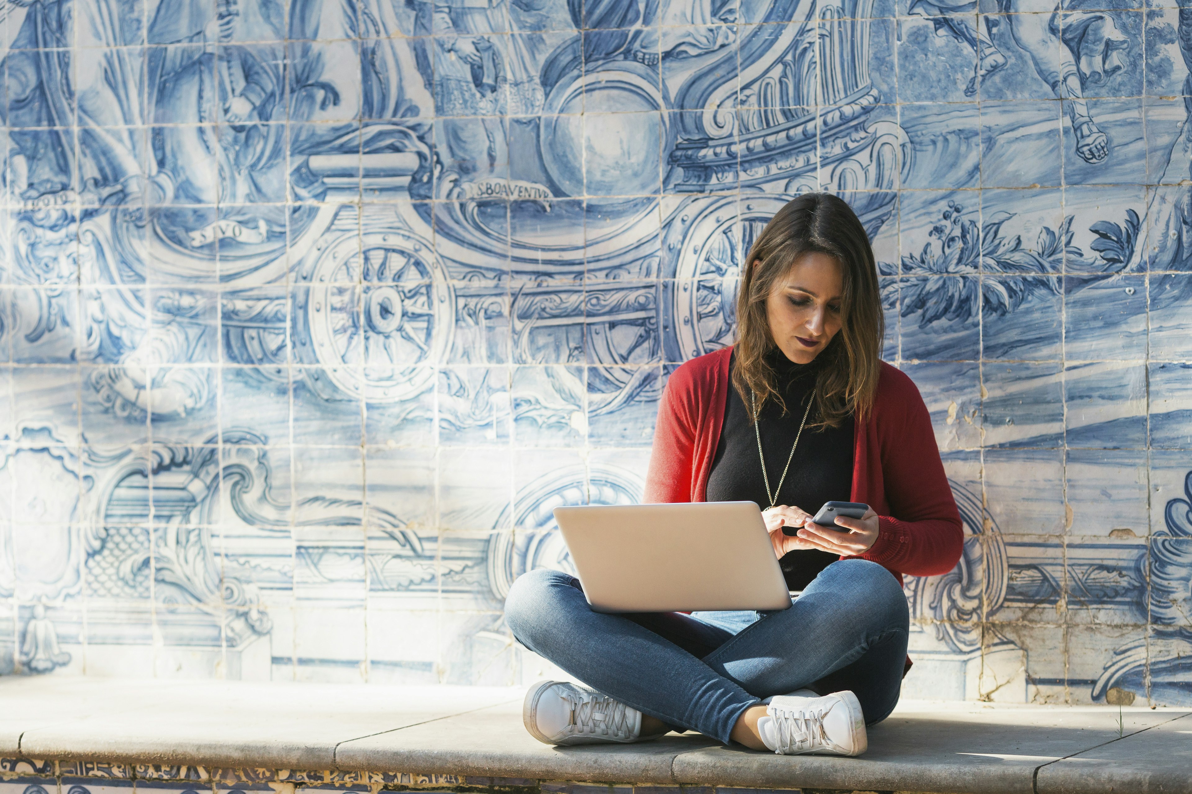 A woman sits cross-legged working on a laptop in front of a blue-tiled wall with traditional designs