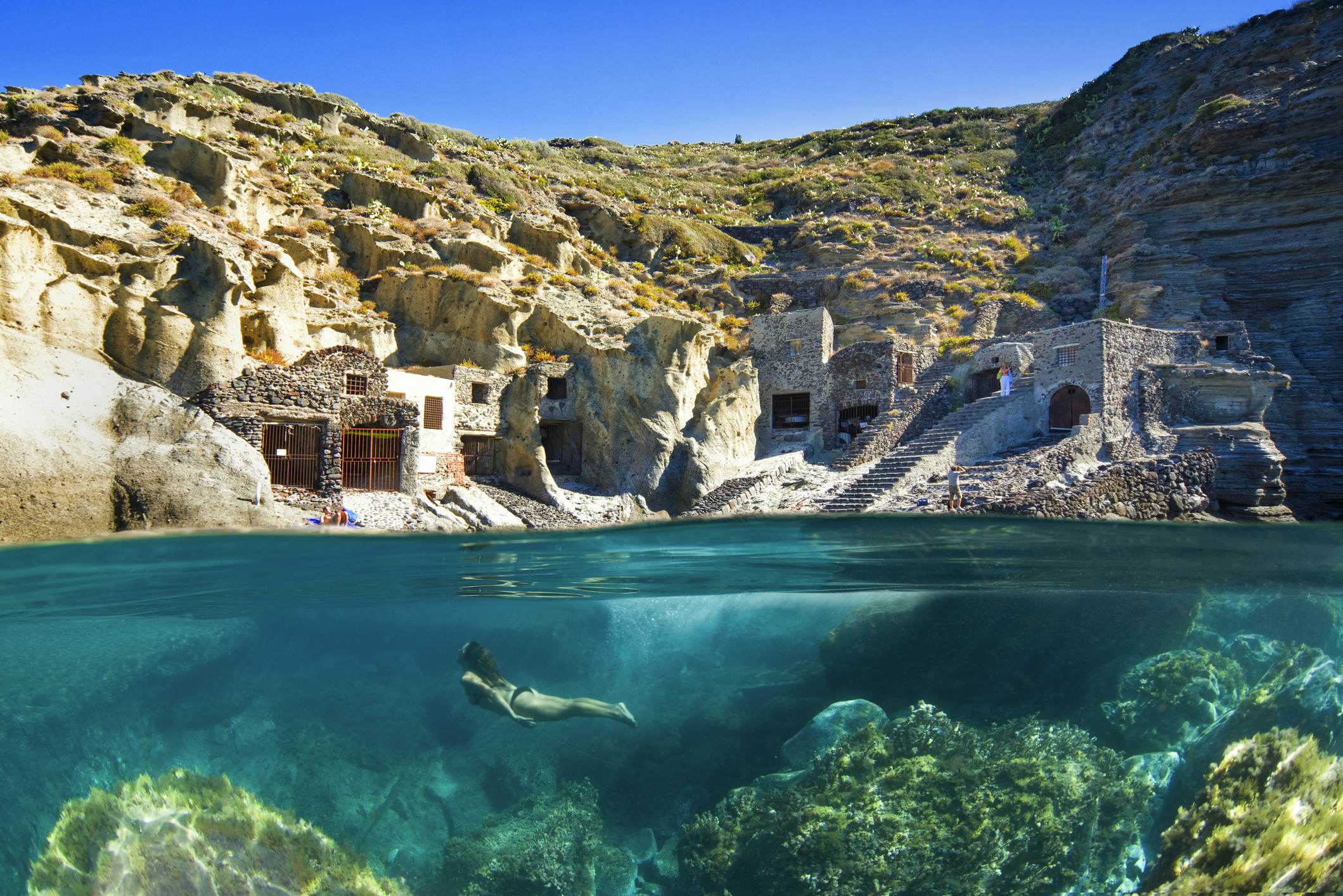 A woman is pictured diving underwater, while the image also shows an above-water scene of buildings carved into a rocky cliff along the shore.