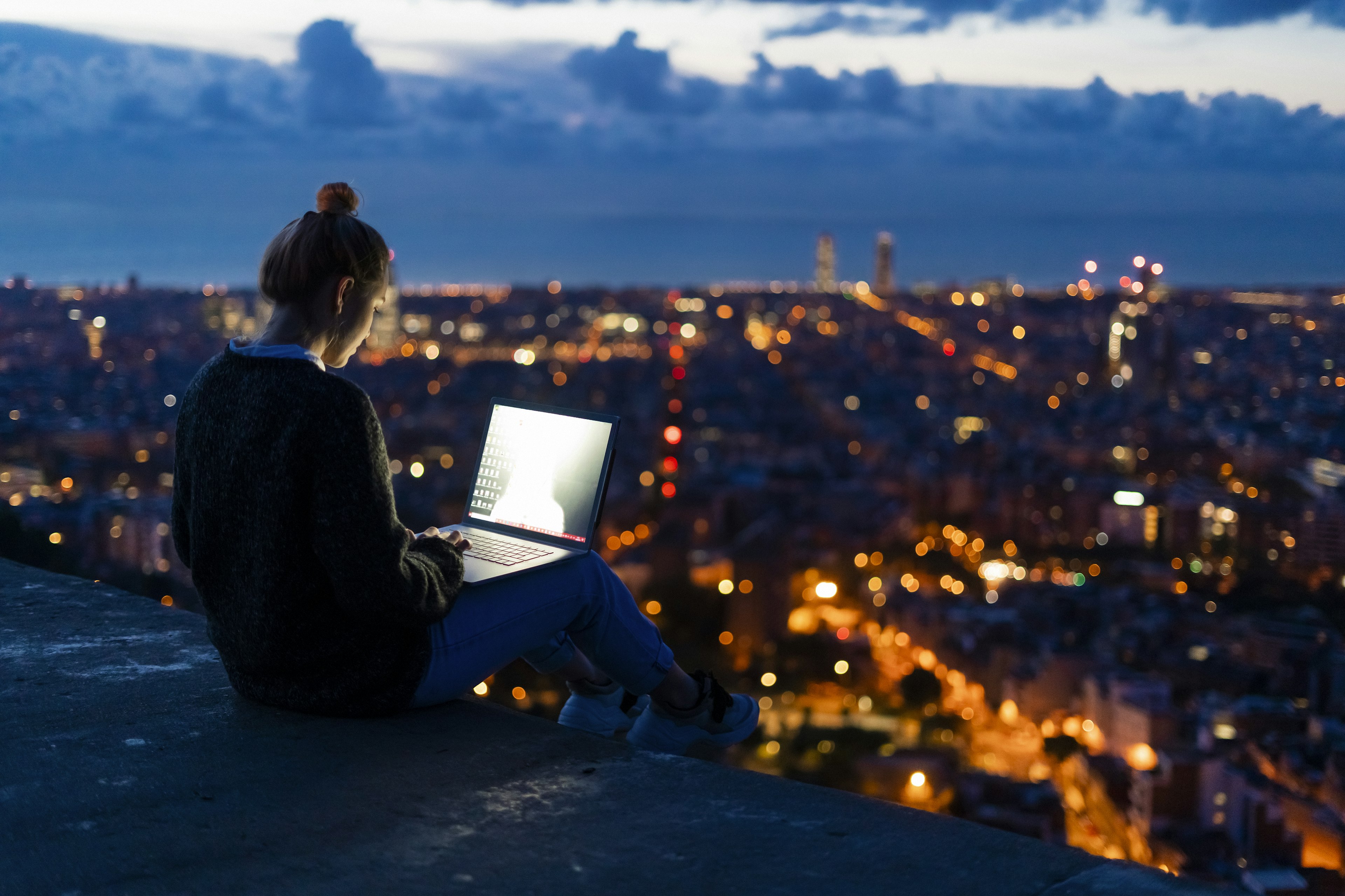 A person sits on a high vantage point during twilight hours, working on a laptop with the city lights sprawling in the background.