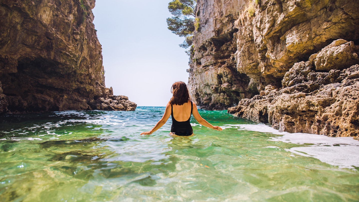 Young adult woman in black swimsuit walking into sea between rocks cliff in Croatia