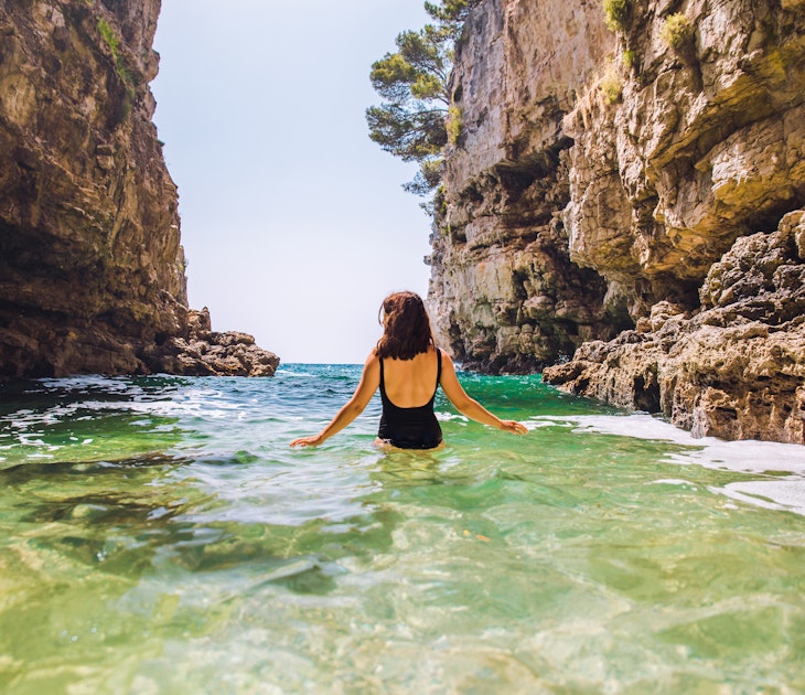 Young adult woman in black swimsuit walking into sea between rocks cliff in Croatia