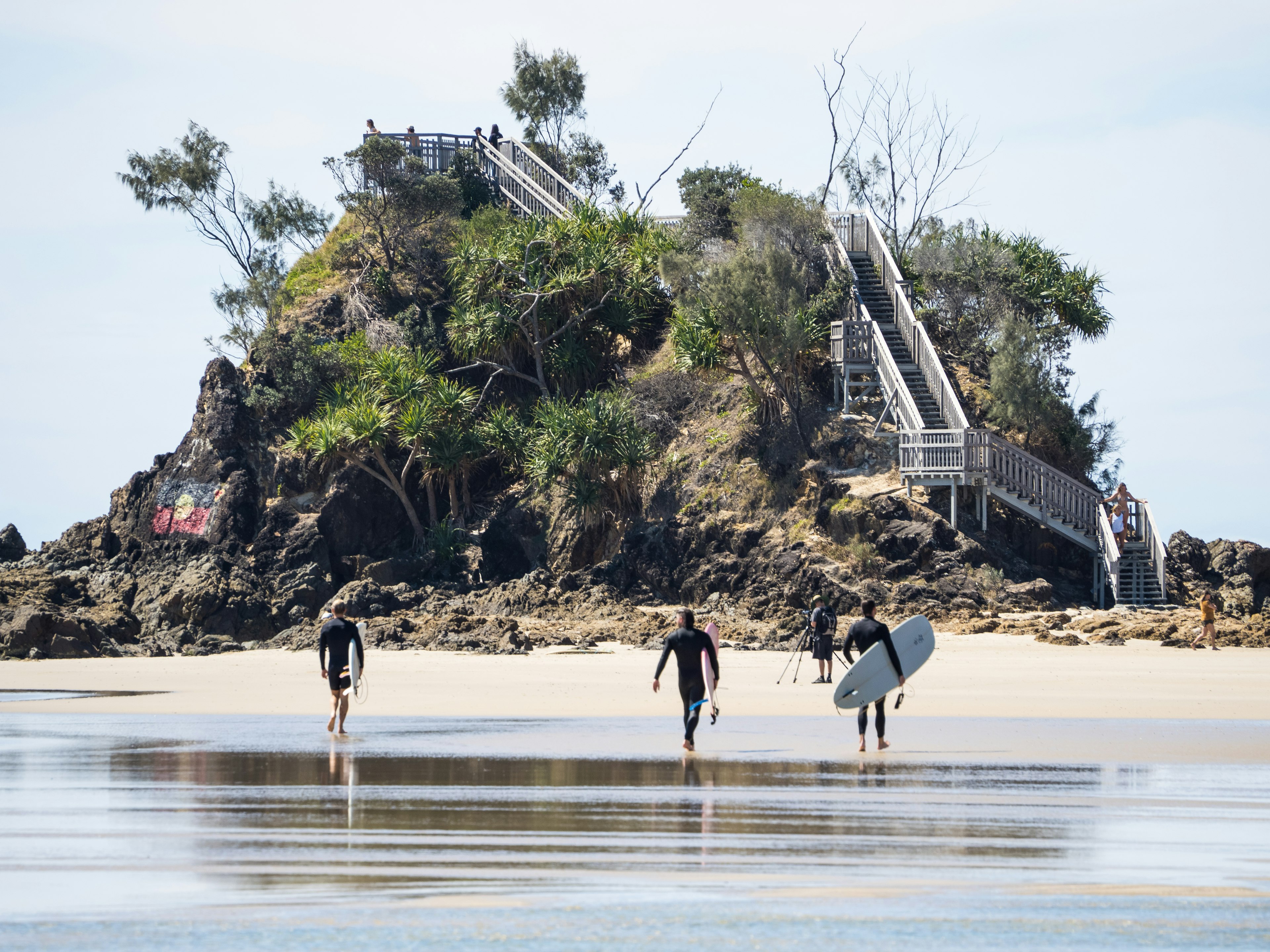 Horizontal landscape photo of male surfers carrying their surfboards along the beach at popular surfing spot, The Pass, Byron Bay, NSW