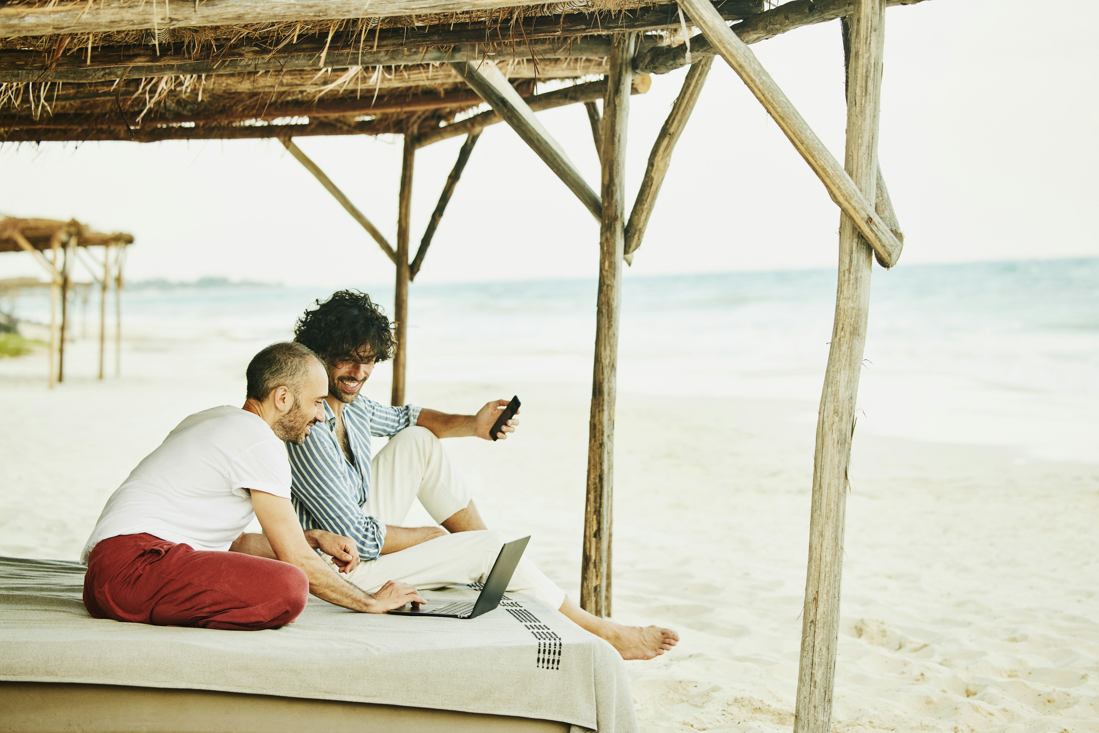 Wide shot of smiling gay couple looking at data laptop while relaxing at beach cabana at tropical resort