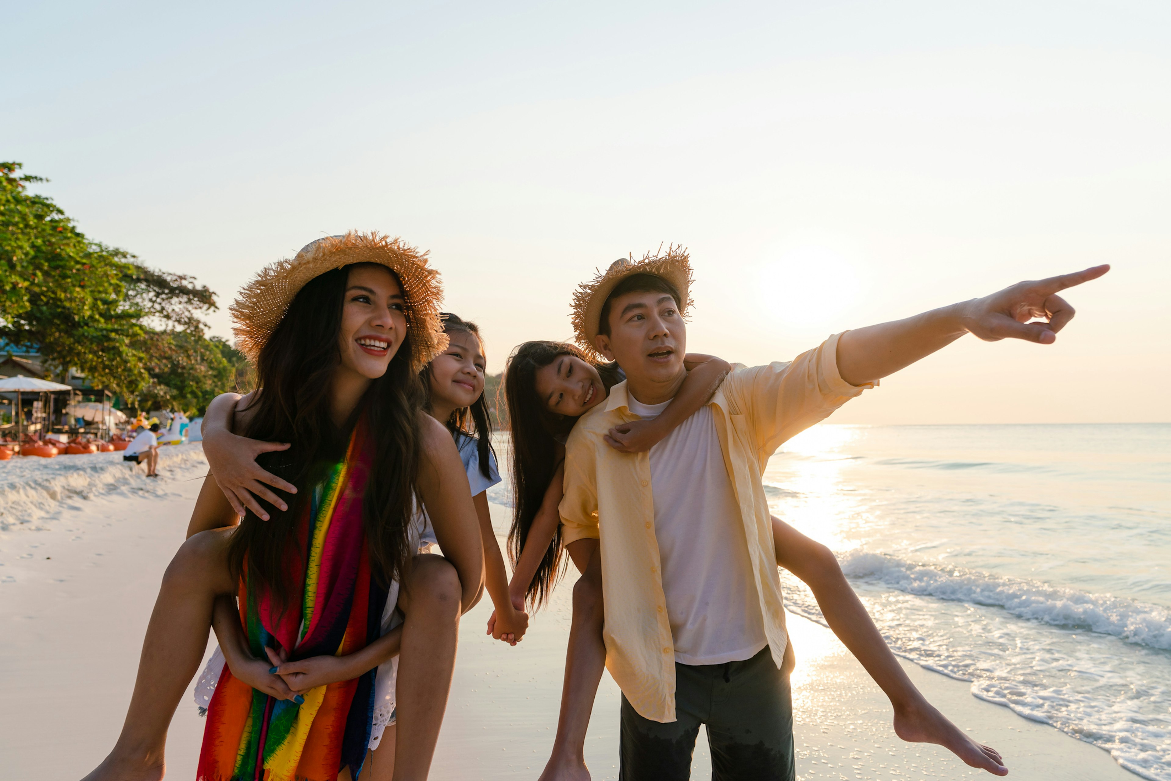 Asian family enjoying summer vacation on the beach in morning with kids on their shoulders and the man pointing to something