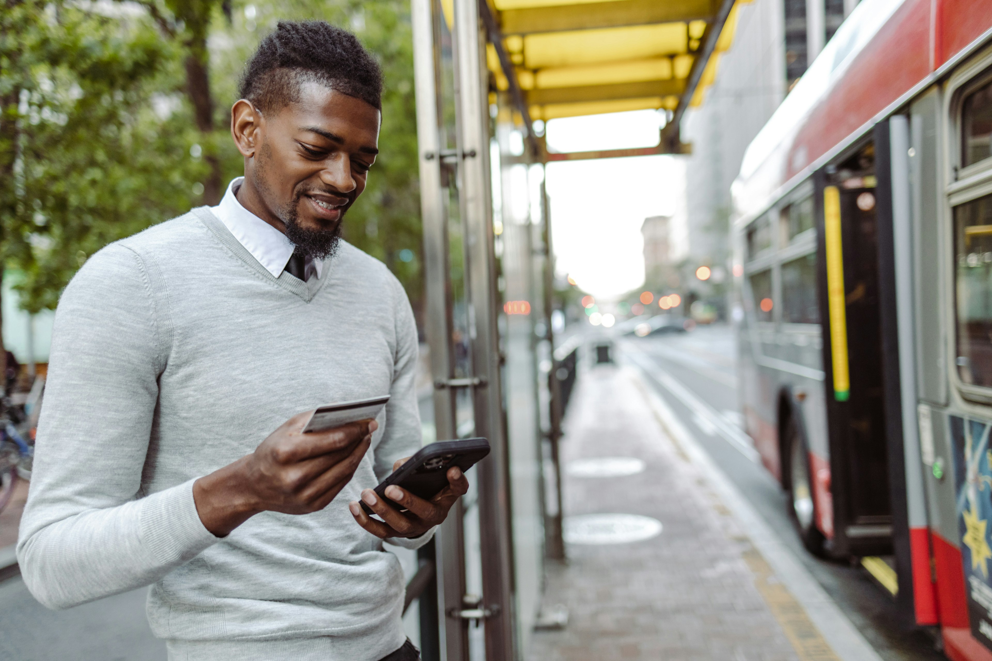 A young man in smart casual attire, looks at his phone at a bus stop