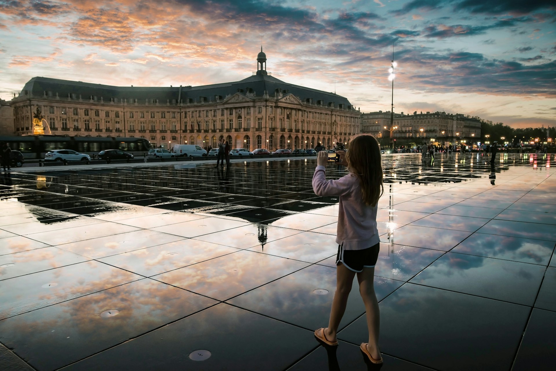 Young girl taking a photo of the famous Bordeaux water mirror with Place de la Bourse in the background at sunset