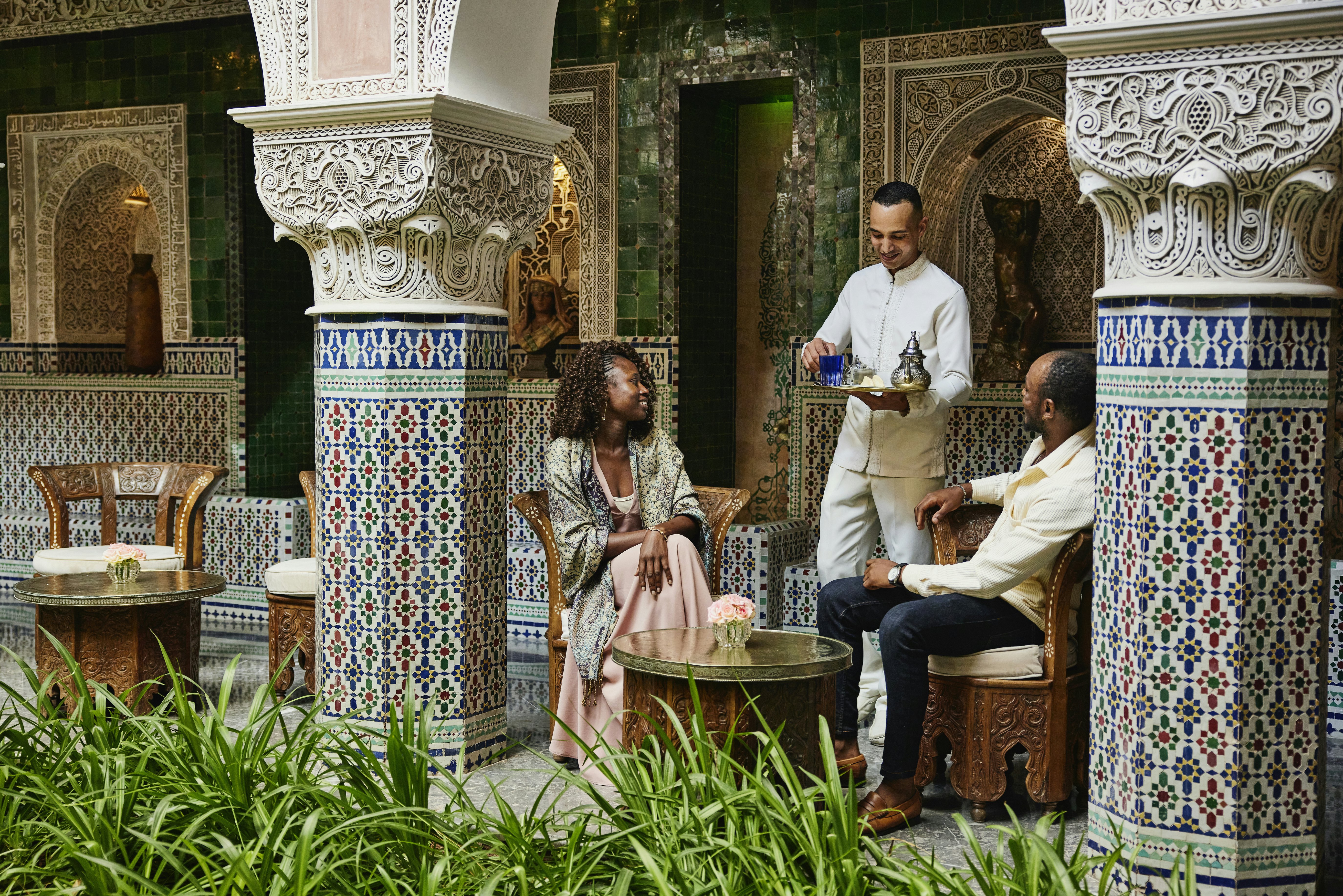 A waiter serves tea to a couple in an ornate Moroccan courtyard with intricate tilework