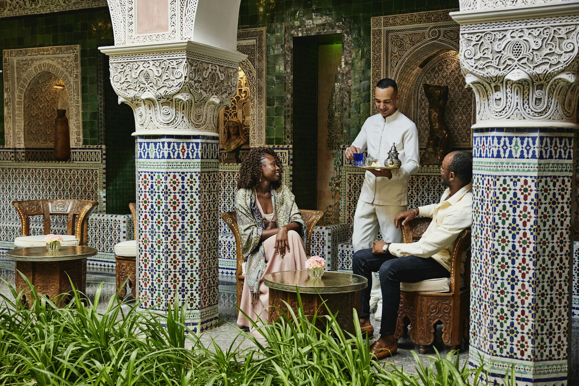 Wide shot of smiling couple being served tea by waiter in courtyard of luxury hotel while on vacation