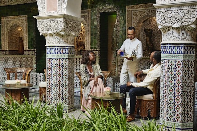 Wide shot of smiling couple being served tea by waiter in courtyard of luxury hotel while on vacation