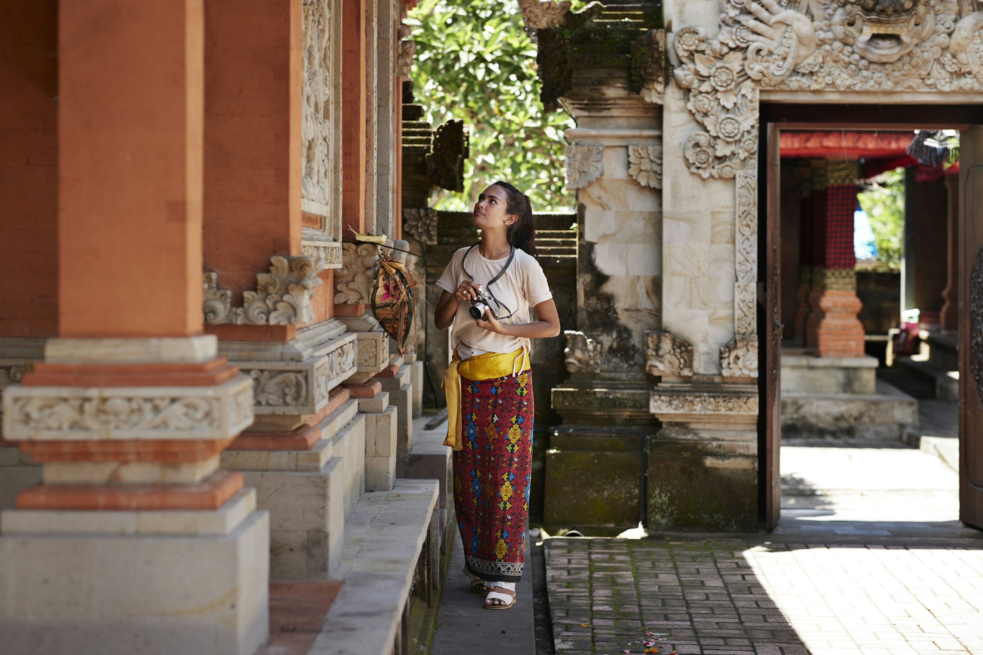 A visitor with a camera and wearing a sarong looks at an architectural detail at a temple courtyard in Southeast Asia