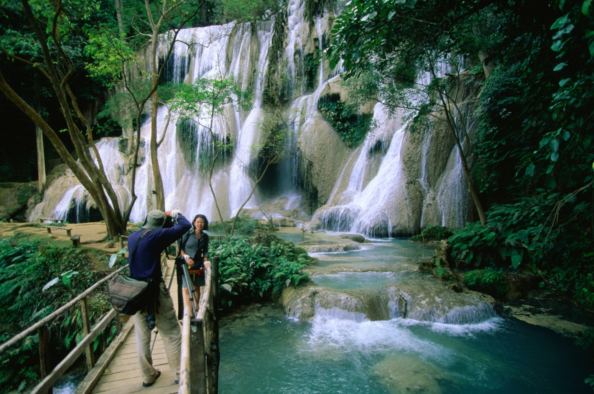 A man takes a photo of his wife in front of some waterfalls at Luang Prabang, Luang Prabang, Laos, South-East Asia