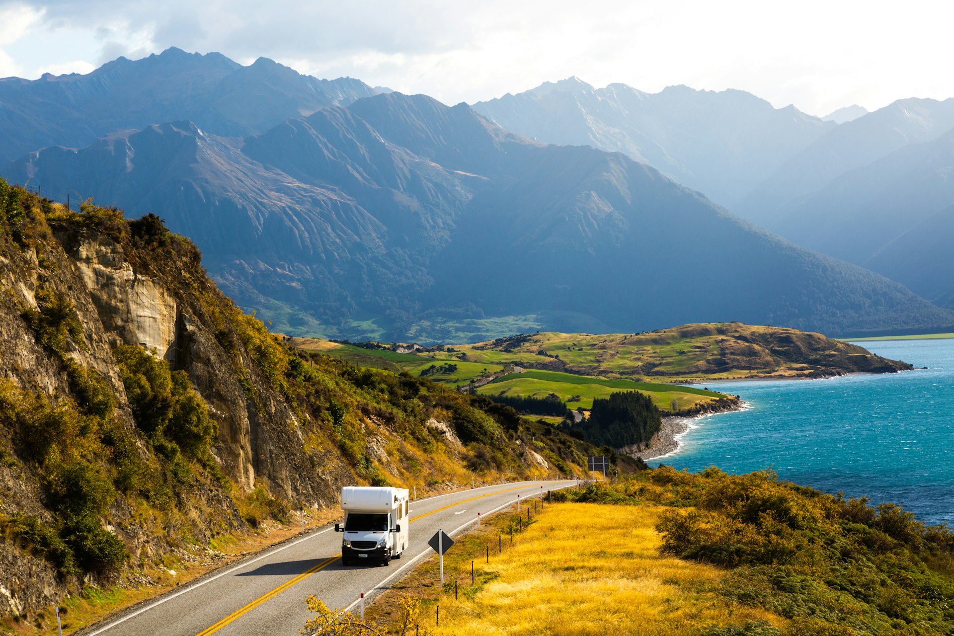 A campervan drives on a New Zealand coastal road with snow-capped mountains in the background