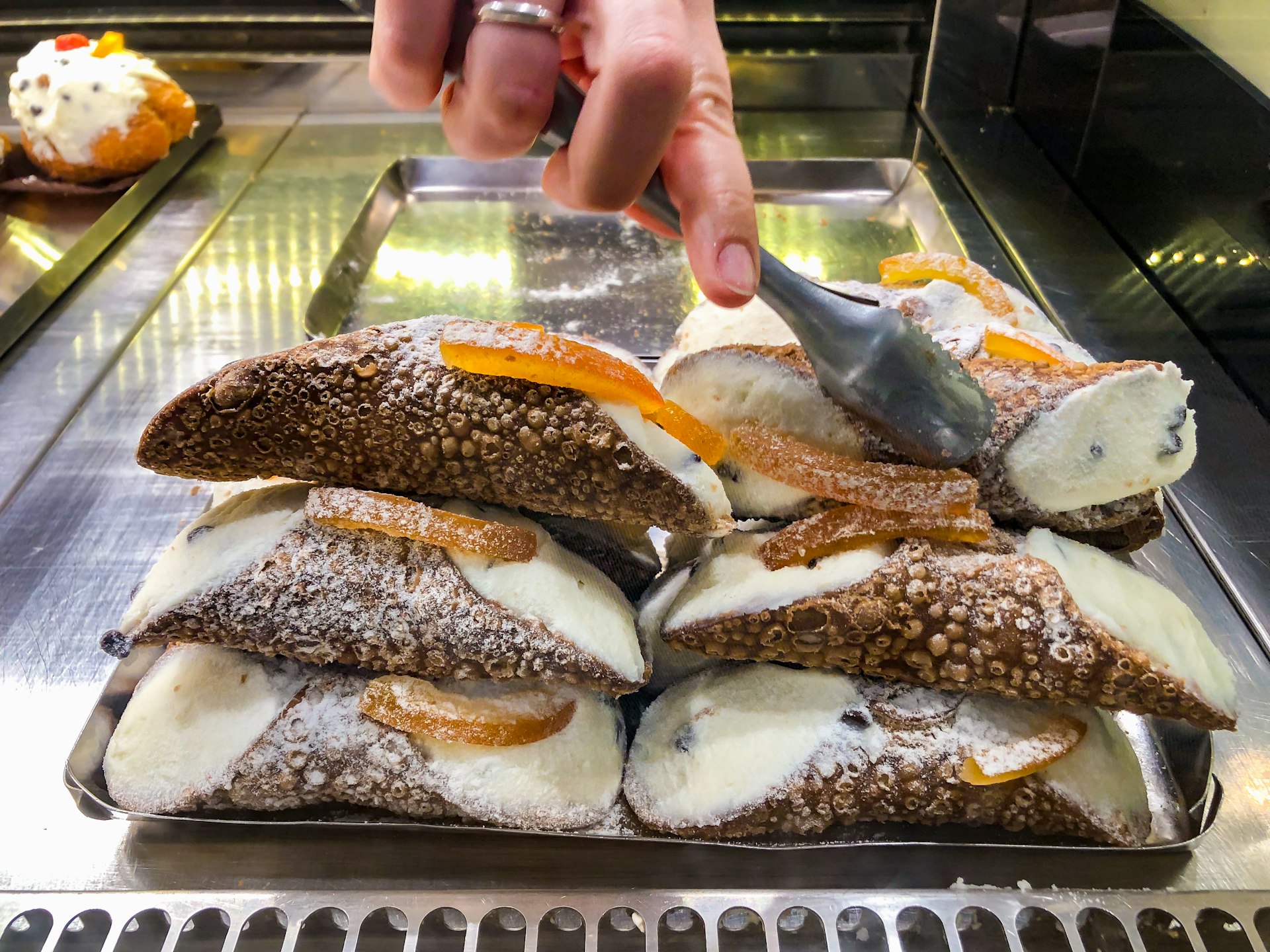  a person in a Sicilian pastry shop serves traditional ricotta cannoli with candied oranges. A pastry chef takes some desserts with pincers
