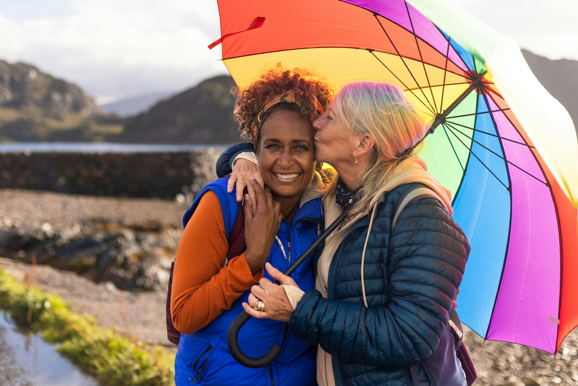 A waist-up shot of a same-sex female couple laughing under a multicoloured umbrella. The woman on the right is kissing her girlfriend on the side of the head. They are standing on the side of Loch Torridon near the village of Diabaig in the west highlands of Scotland.