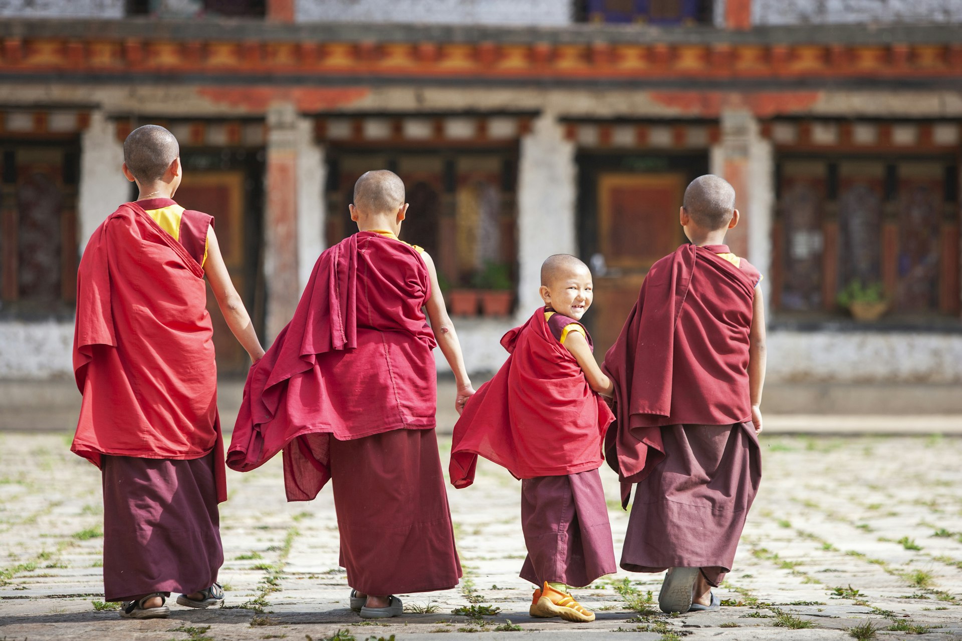 Four novice monks wearing red robes walk through a courtyard, with one giving a cheeky smile at the camera