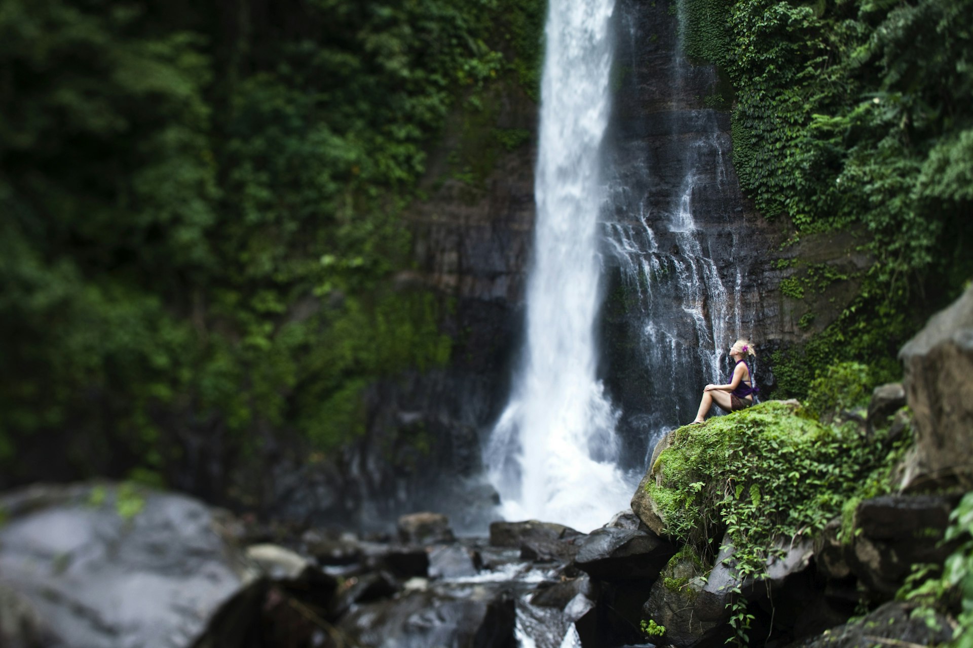 A woman sits close to a waterfall in Bali which is surrounded by verdent green forest