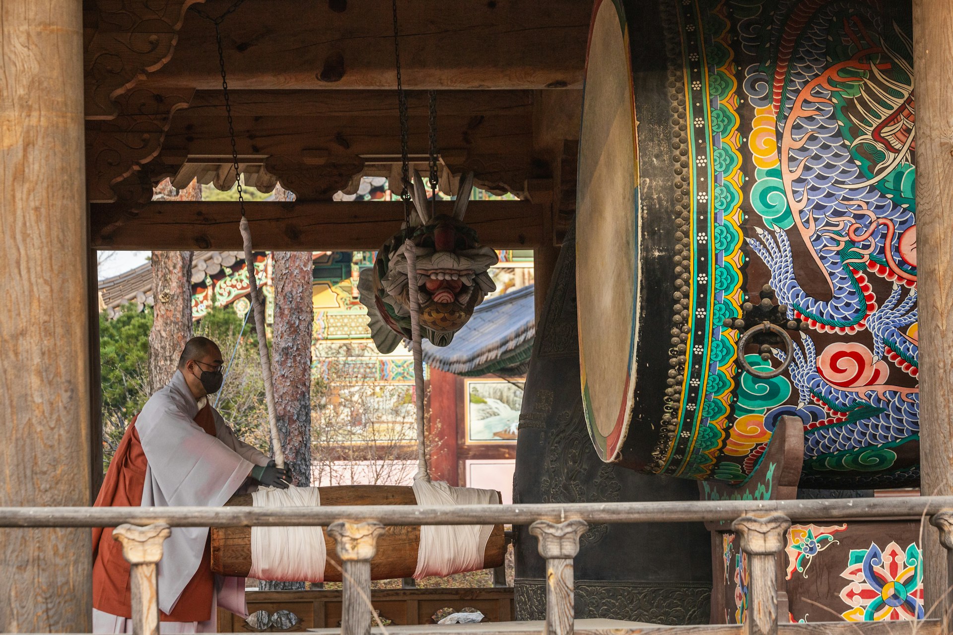 A Buddhist monk in traditional attire and a face mask stands inside a temple pavilion, striking a large, ornate hanging drum with a mallet. The drum is adorned with intricate patterns, and beside it, part of a colorful mural with a dragon motif is visible, showcasing the rich artistic heritage of the temple. The pavilion's architecture, with its wooden beams and decorative elements, reflects the traditional Korean style. The atmosphere is one of solemnity and cultural significance.