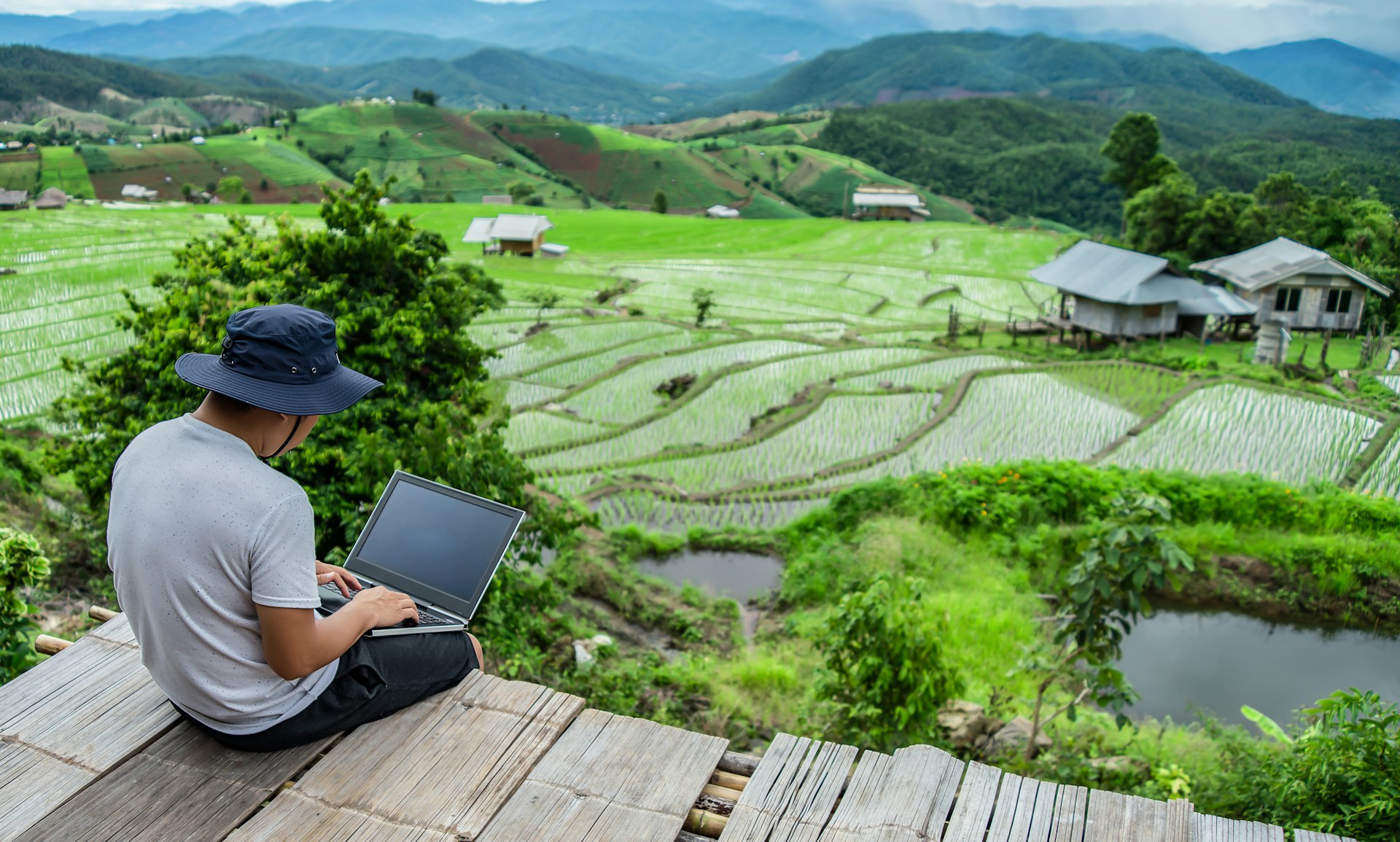 A person is typing on a laptop while sitting outside, overlooking a lush green landscape.