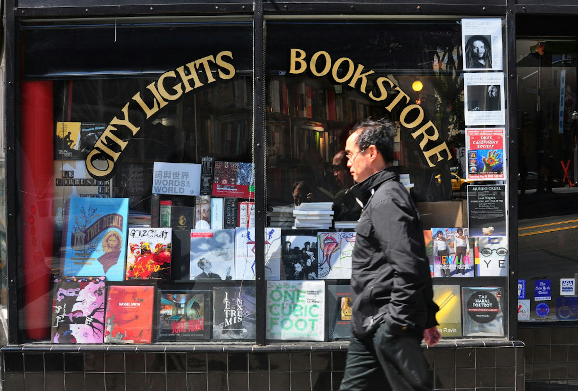 A man walks past the storefront of City Lights Bookstore. The store's window displays an array of colorful book covers and posters, reflecting a vibrant literary culture. The iconic City Lights sign is prominently featured above the window.