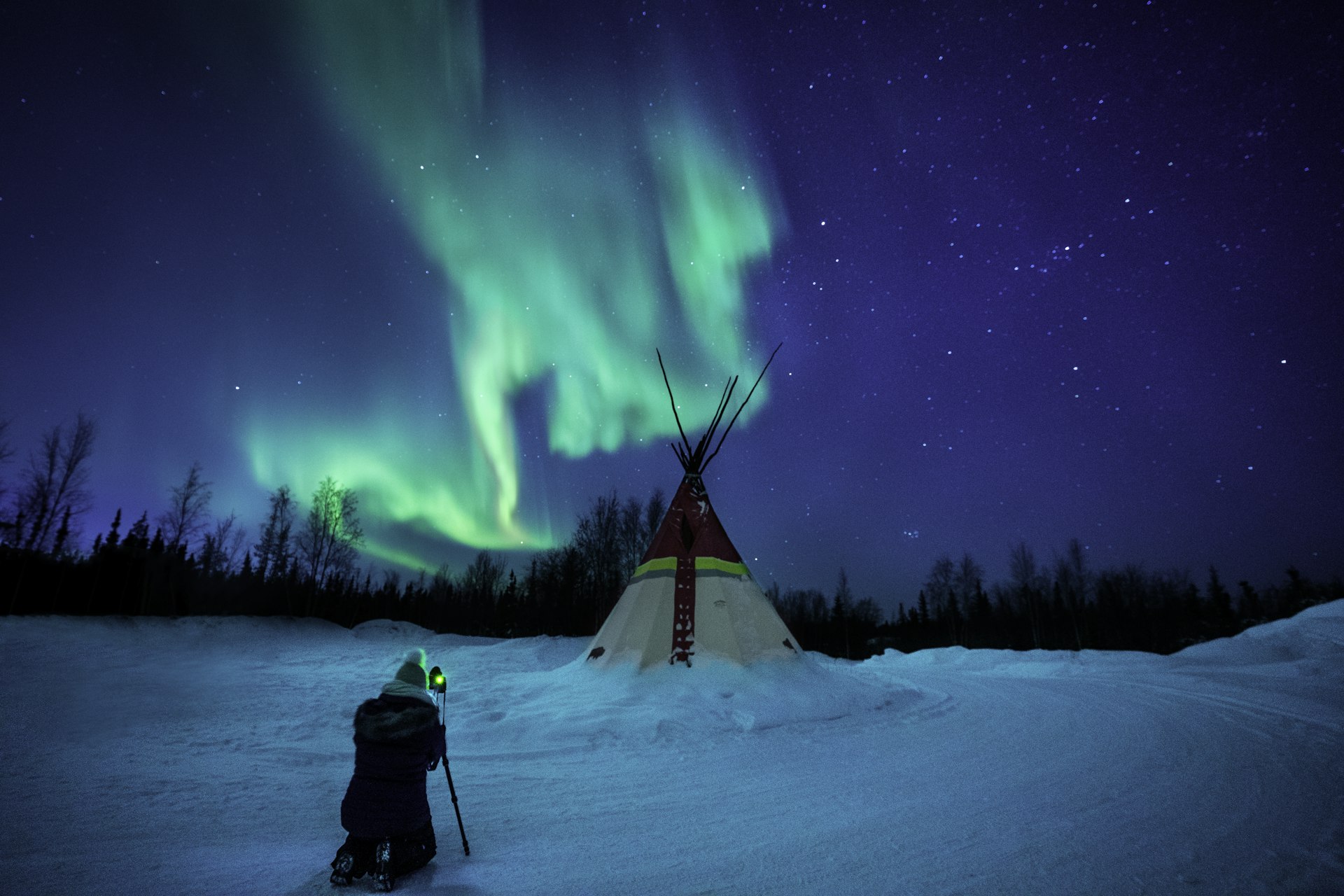 GettyA photographer in winter clothing takes a photo of a brightly lit traditional teepee under the green glow of the Northern Lights in a snowy landscape