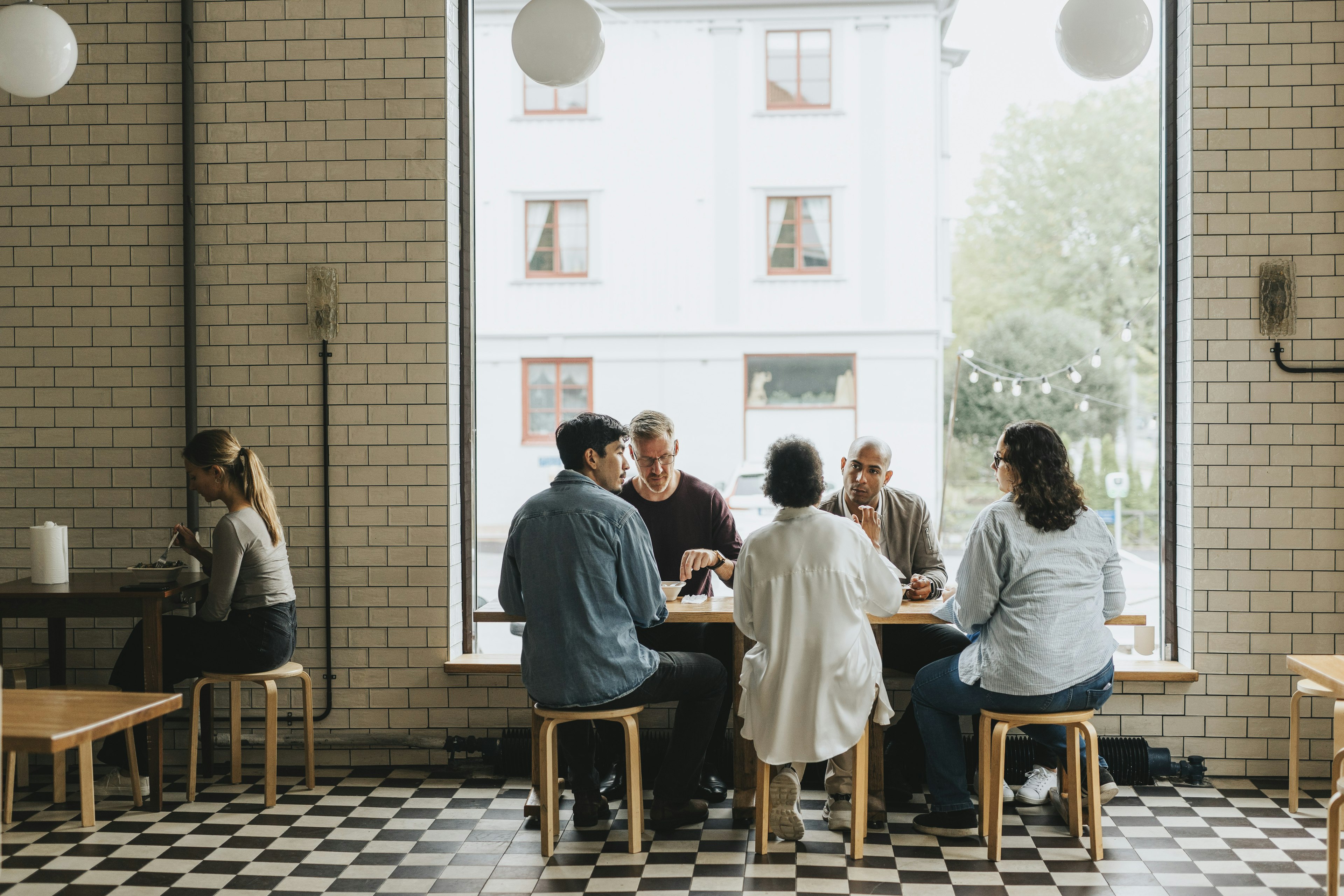 A group of people engaged in conversation at a communal table inside a modern cafe with large windows, black and white checkered flooring, and an unoccupied table in the foreground