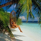 Woman in cane chair hanging from palm over beach, Ihuru, Maldives