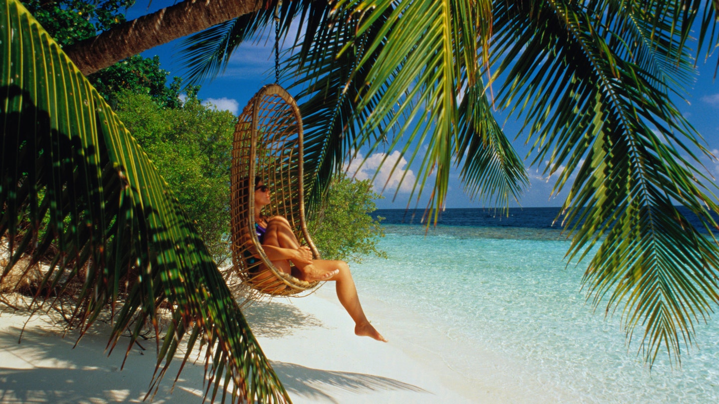 Woman in cane chair hanging from palm over beach, Ihuru, Maldives